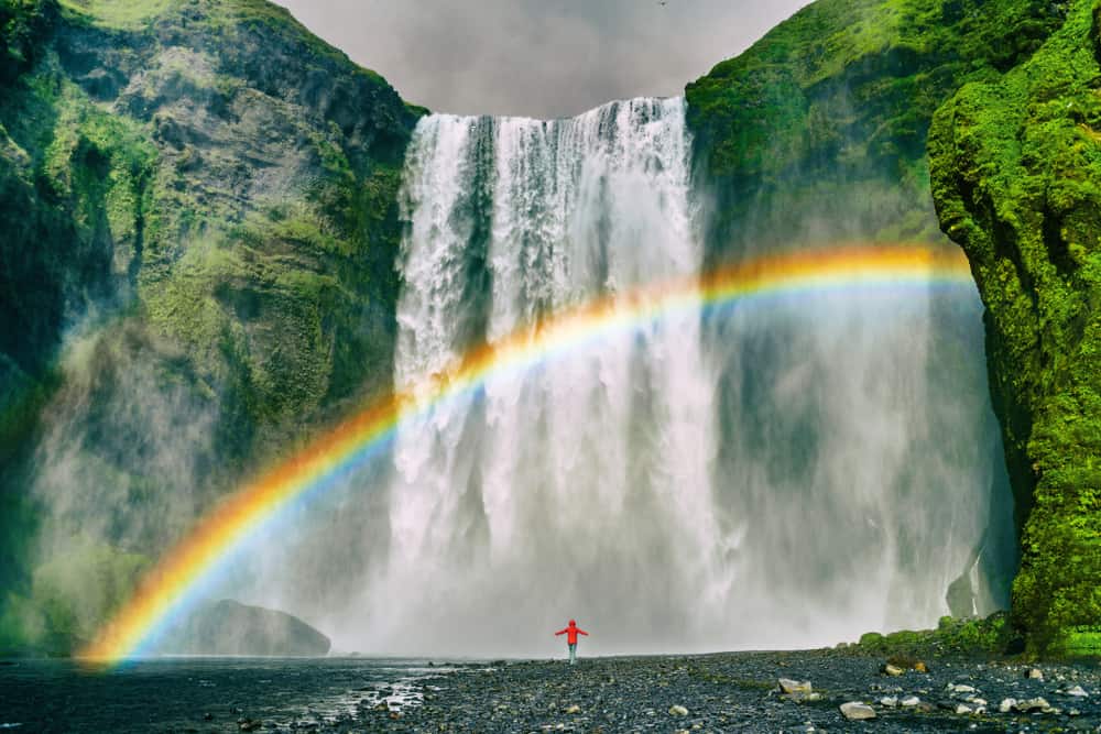 A figure in red stands under a rainbow over Skogafoss waterfall in South Iceland.