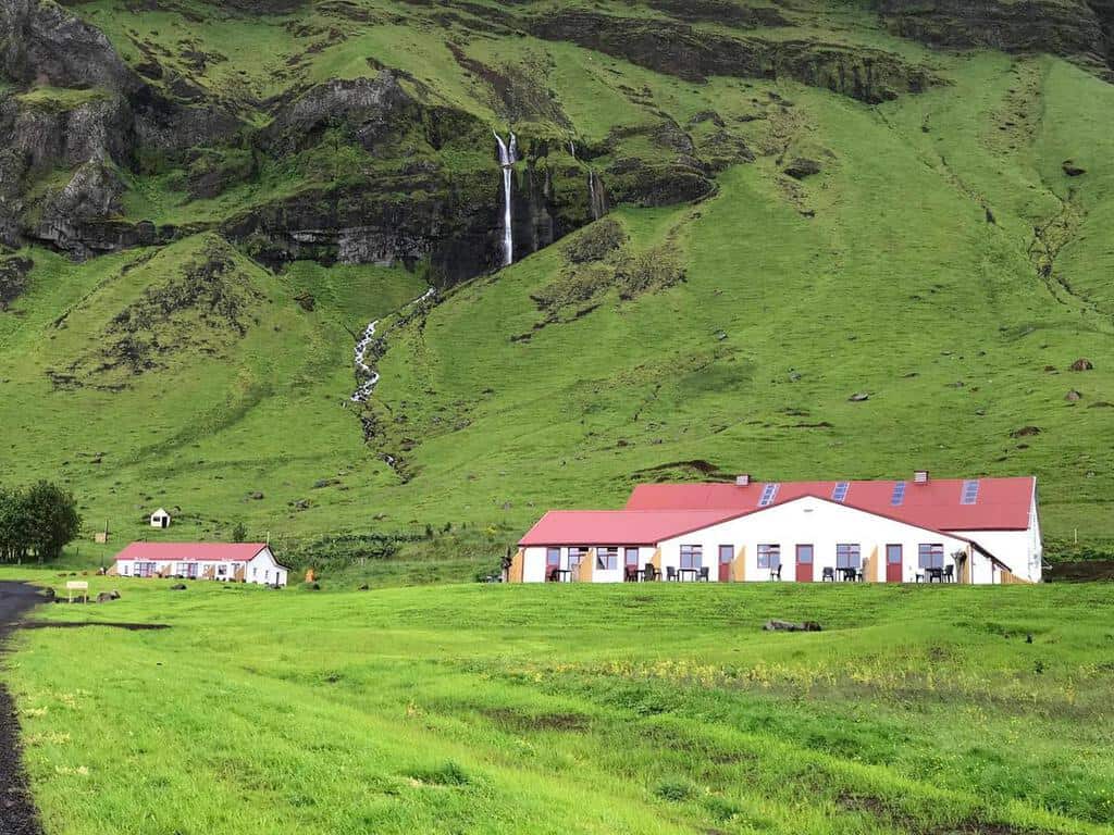 white buildings with red roofs and a field with a large mountain and a waterfall in the background