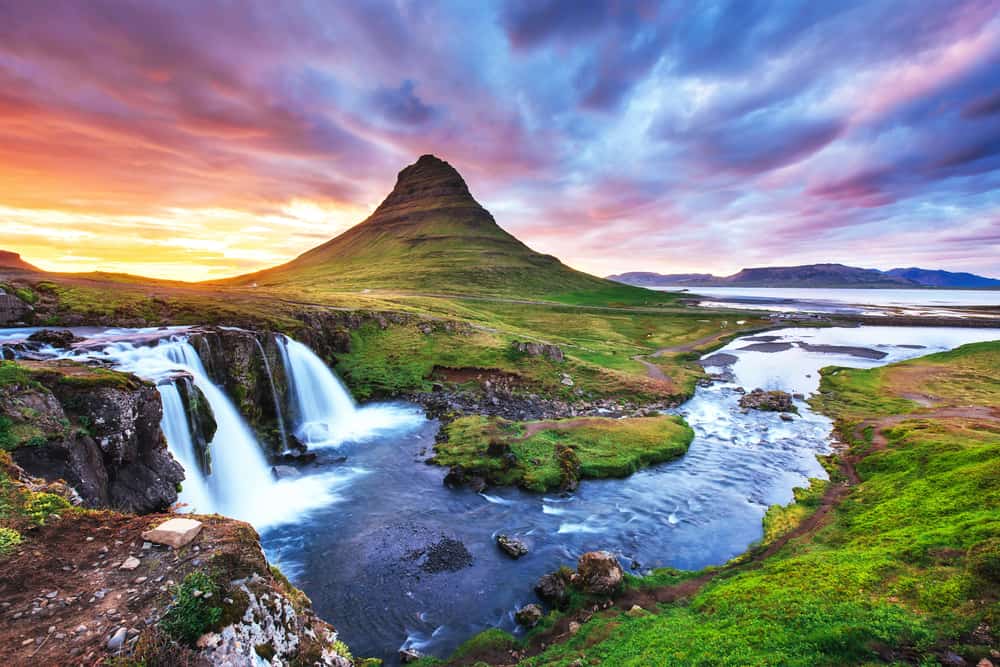 A double waterfall flowing in to river below with a large green mountain in the background at sunset