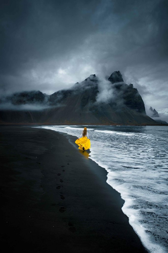 A woman in yellow on black sand beach in front of vestrahorn mountain, one of the best things to do in Iceland.