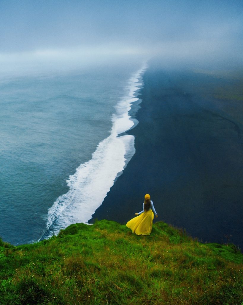 A woman in yellow overlooking black sand beach from viewpoint.