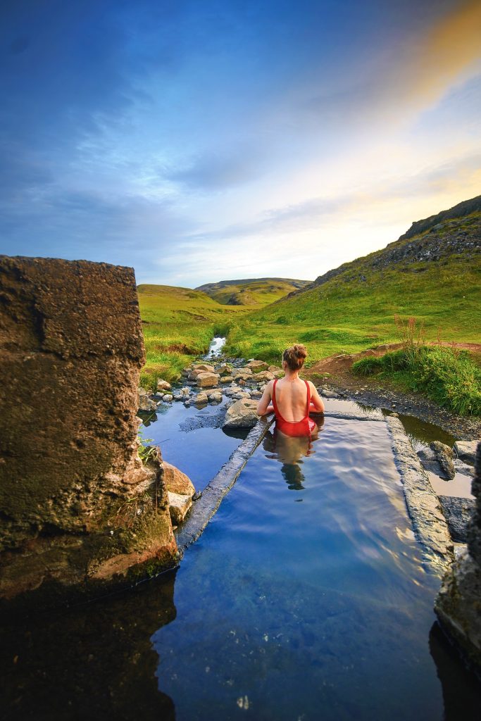 Woman in red swim suit stands in Hrunalaug Hot Springs at sunset.