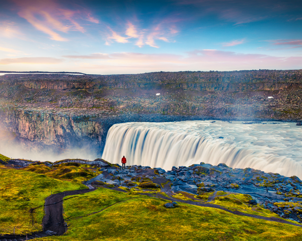 a person in a red jacket standing in front of the mighty Dettifoss waterfall