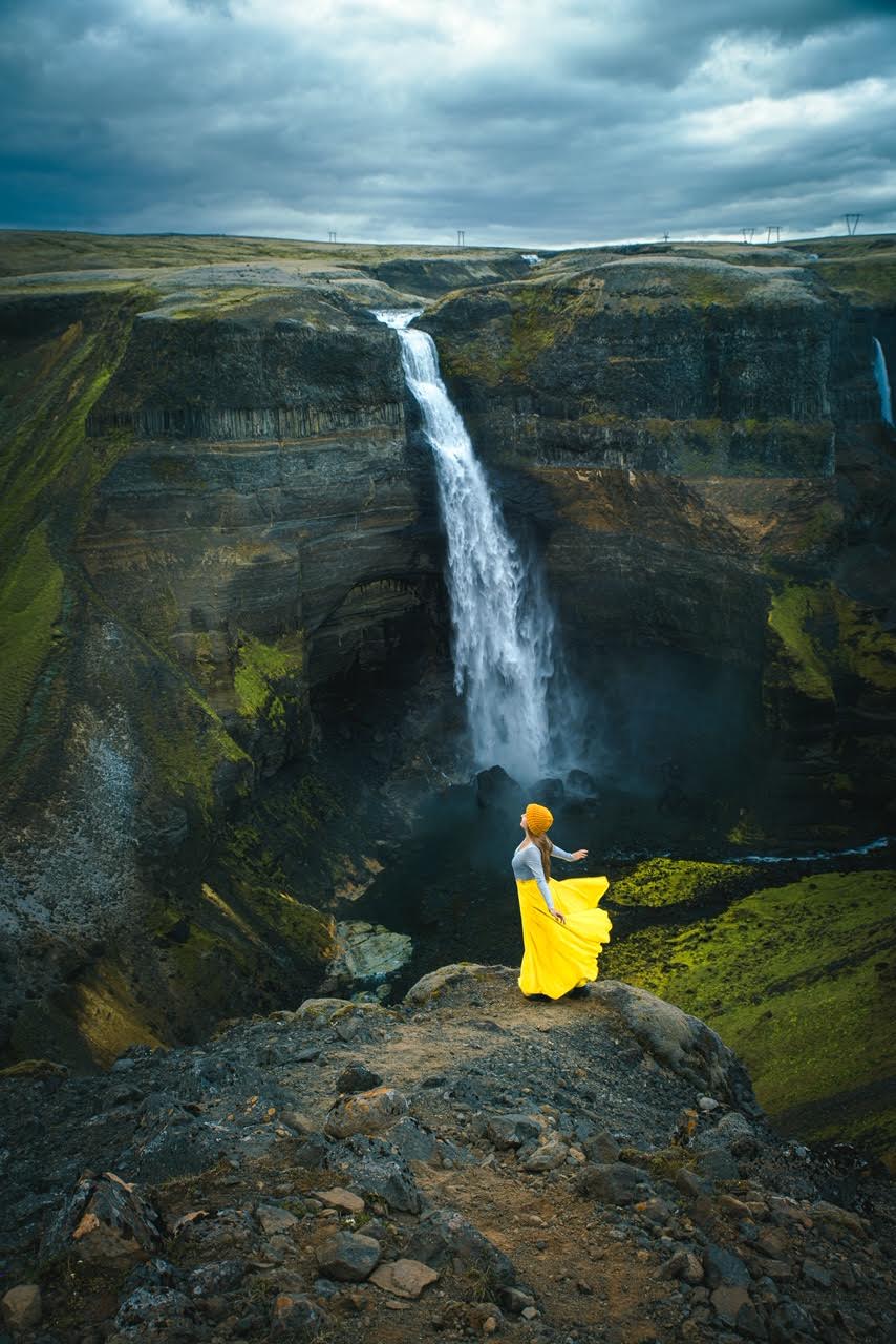 Woman standing with arms out wearing a long flowy yellow skirt and a big waterfall in the background