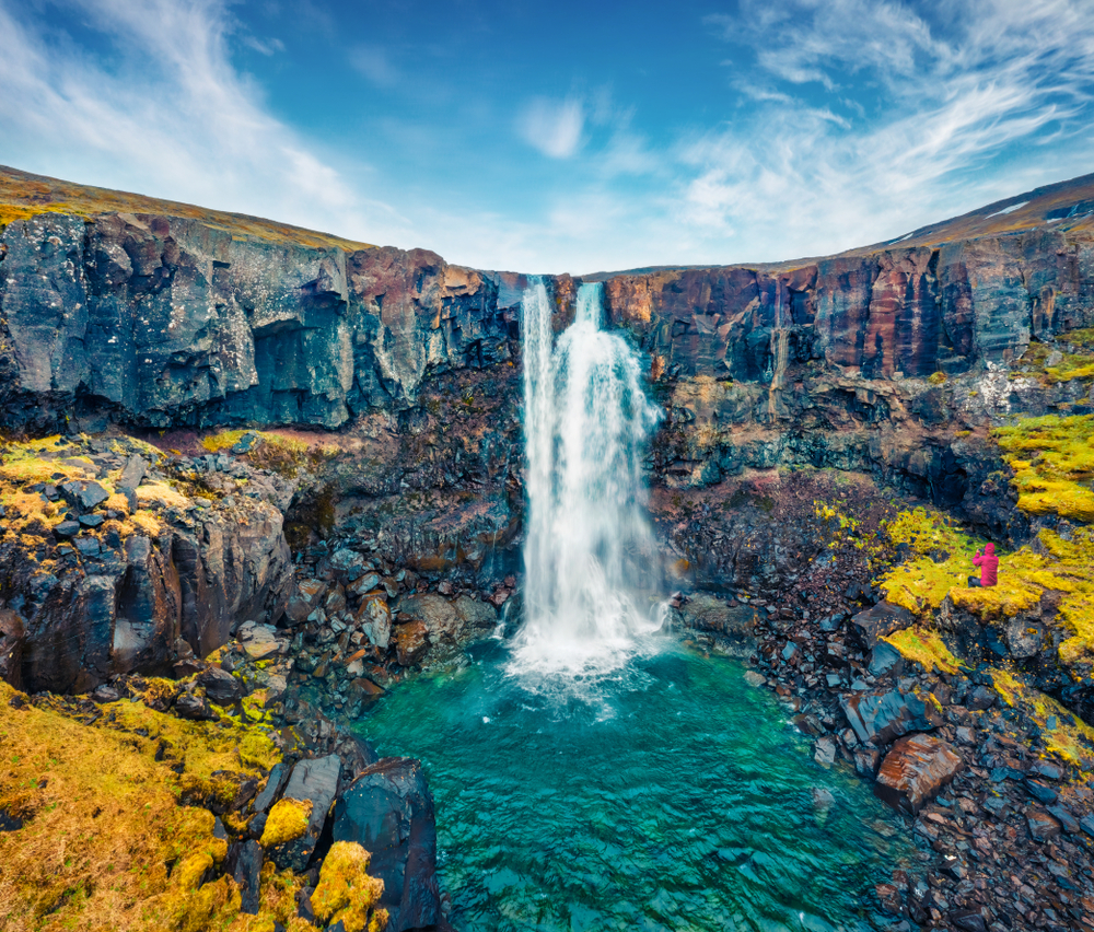 a person small in the frame in a pink jacket taking a picture of a waterfall