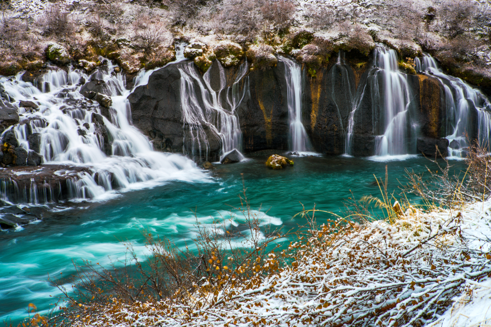 multiple small waterfalls flowing into blue river below with snow on the ground