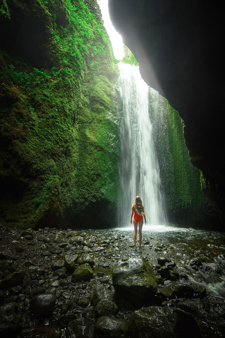 Women standing in a red swimsuit in a cave looking up at a waterfall 