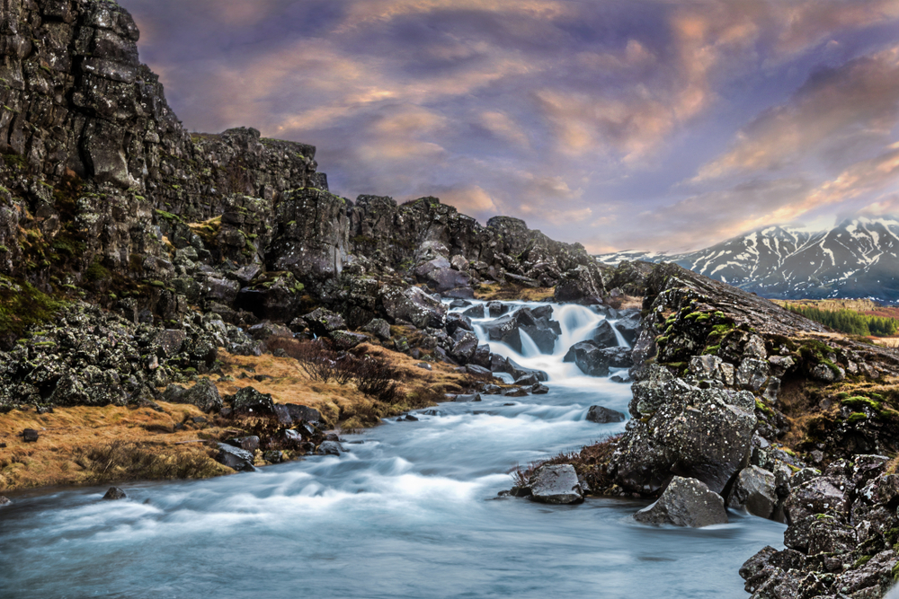 a small waterfall flowing into a raging river surrounded by large rocks at sunset