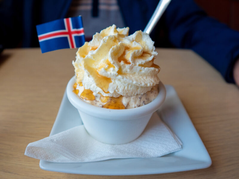 A bowl of ice cream with whip cream and a caramel drizzle sits on a table with a little Iceland flag for decoration.