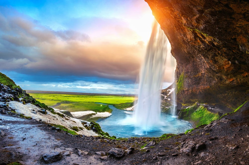 View from behind Seljalandsfoss Waterfall with melting snow and grass turning green for April, the best time to visit Iceland to see the seasons change.