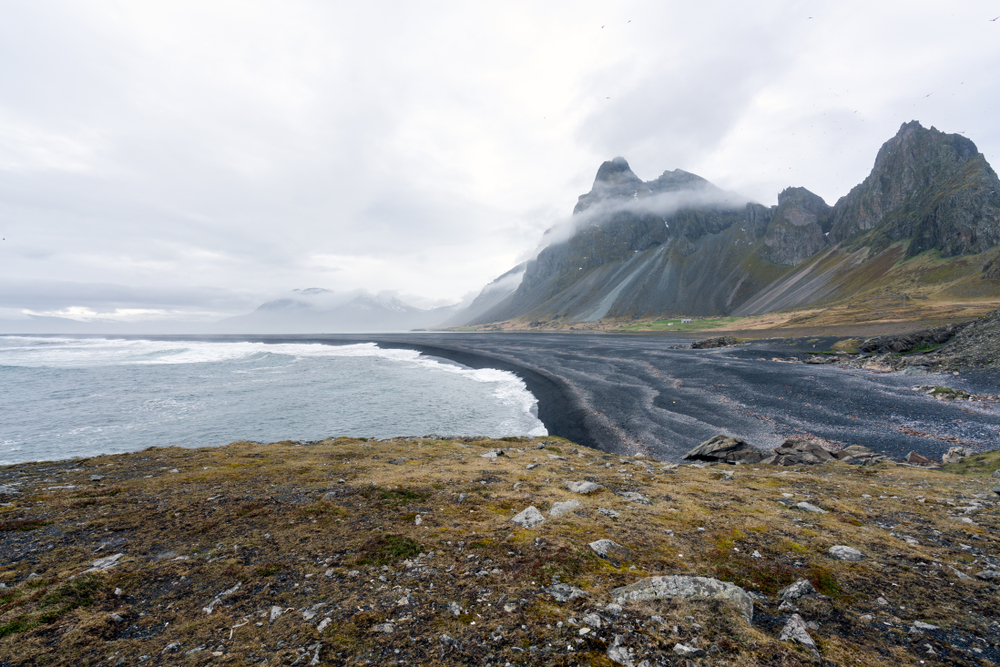 photo of the black pebble beach in Hvalnes national park. There are mountains in the background with fog over them