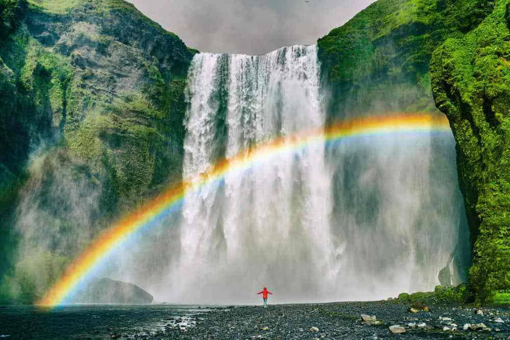 A person standing in a red jacket looking at a majestic waterfall with a rainbow in the mist