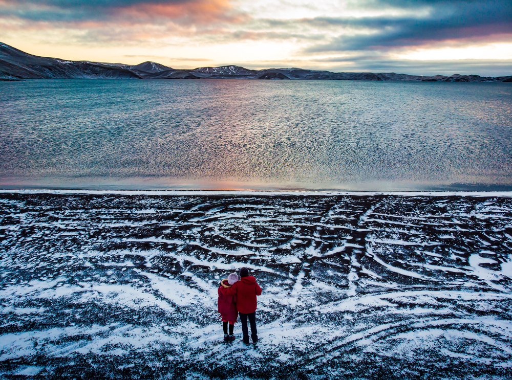 2 people in red jackets standing at Kleifarvatn lake with snow on the ground 