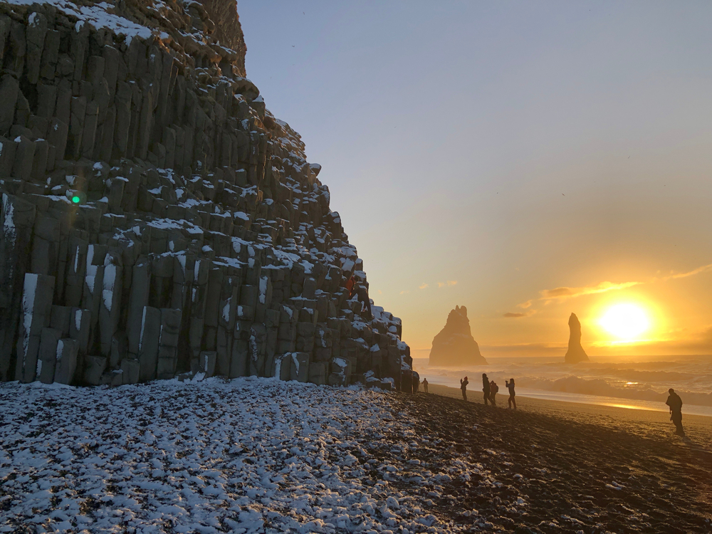 people walking on Reynisfjara black sand beach in Iceland in the winter at sunrise with snow on the ground