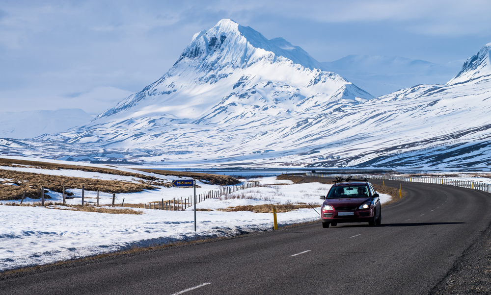 a single red car driving on a paved road with snow mountains in the background