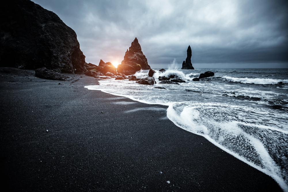 Reynisfjara Black Sand Beach with the sun peaking out from the horizon on a cloudy day.