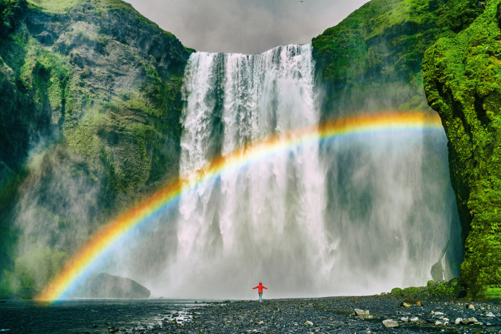 Figure standing under a rainbow at Skogafoss Waterfall.