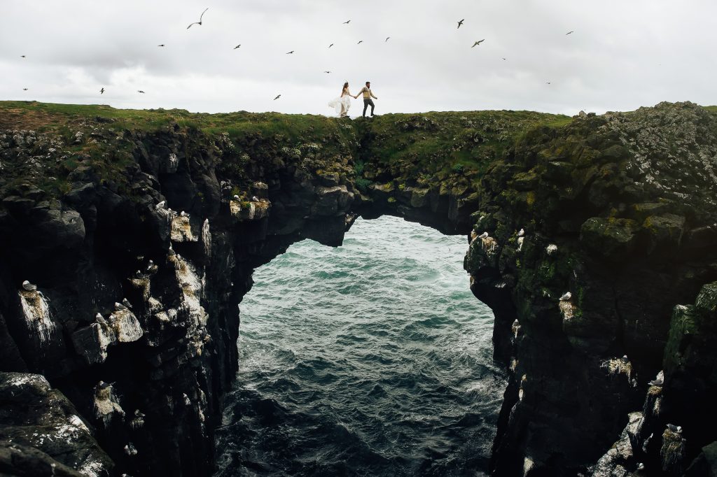 couple crossing rock arch bridge in ocean together Iceland wedding