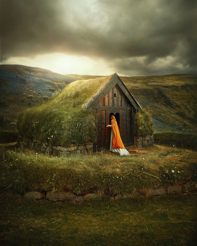 woman in white dress and orange coat peering into grass roof building Iceland wedding