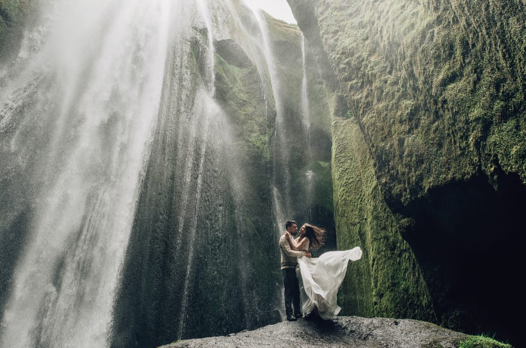 couple in wedding attire standing in front of waterfall, hair and dress flowering Iceland wedding