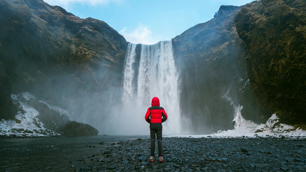 man standing in front of skogafoss waterfall in iceland wearing a red jacket 