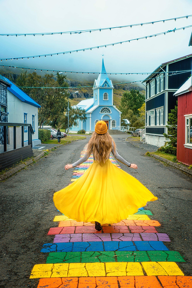 Woman in yellow dress walking in  towards a blue church on a rainbow path one of the best hidden gems in Iceland