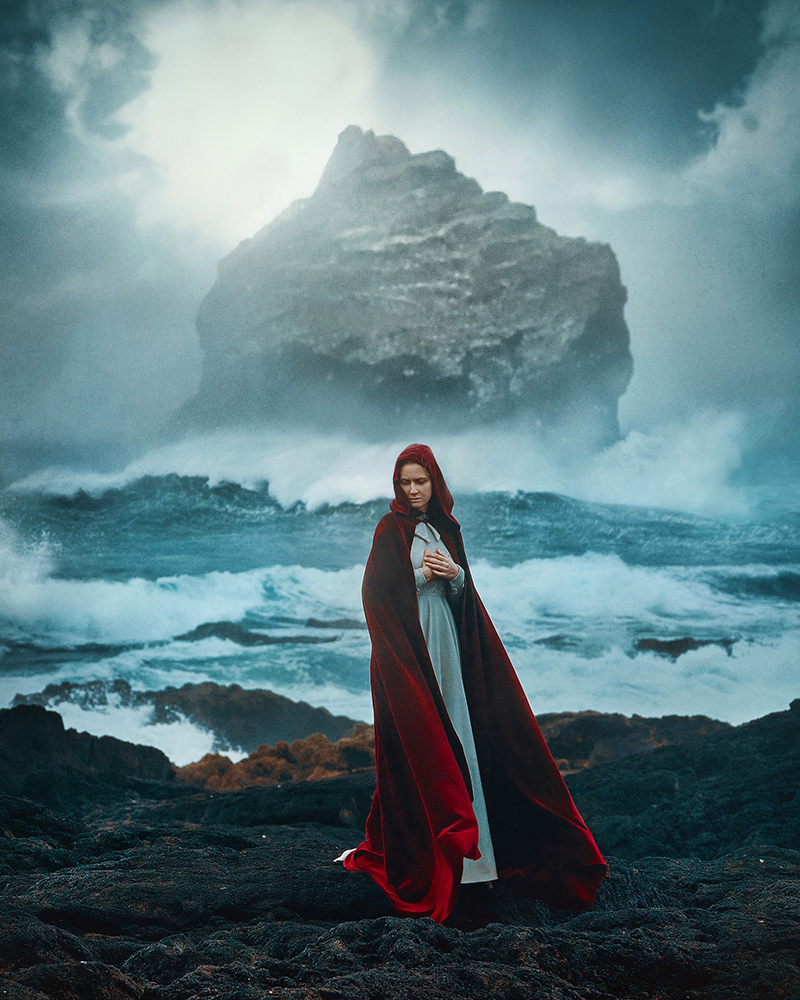 Woman in long, red cape standing in front of ocean and large rock in Iceland