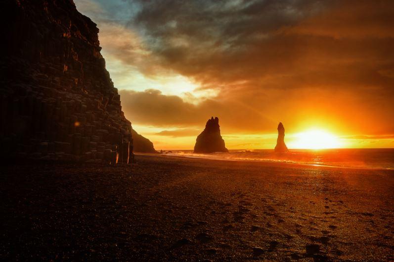 sunrise on a black sand beach with basalt rocks on the left and distant sea stacks in the ocean