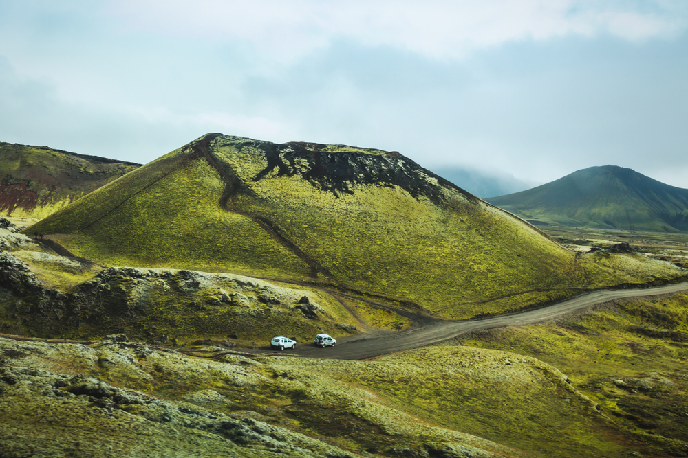 2 white cars driving on a dirt road surrounded by green hills on a moody day in iceland in spring