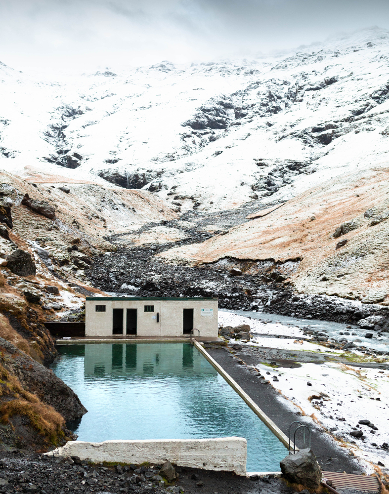 A man made swimming pool hidden in a valley in Iceland. there is snow on the ground, large rocks all around it, and in the distance the slope of a snow covered mountain. At the end of the pool is a small building that is cream with black doors. 