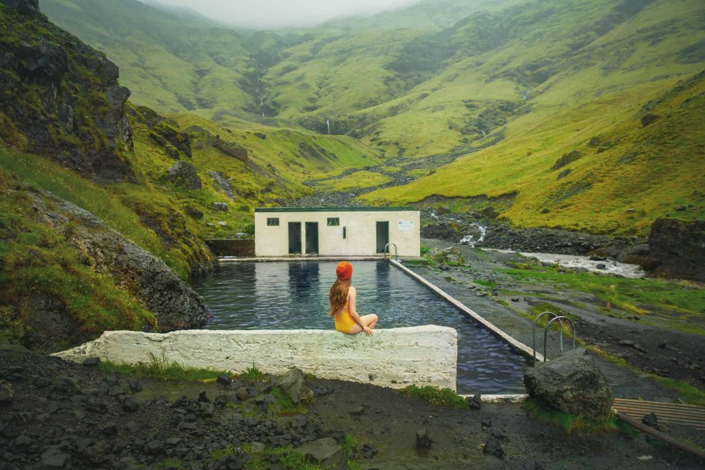 Woman with long hair sitting on a half wall above Seljavallalaug pool wearing a yellow beanie and yellow swimsuit, looking towards the pool, the changing rooms and distant mountains 