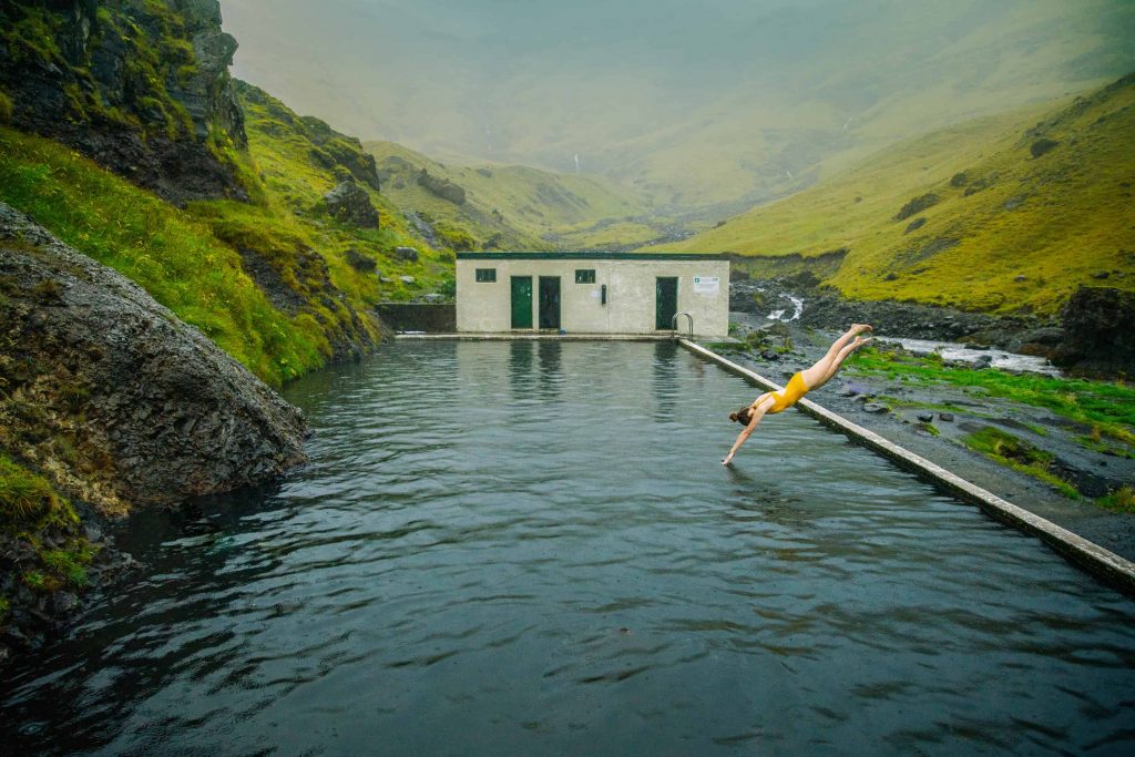 woman in yellow bathing suit diving into Seljavallalaug pool in iceland on a cloudy day