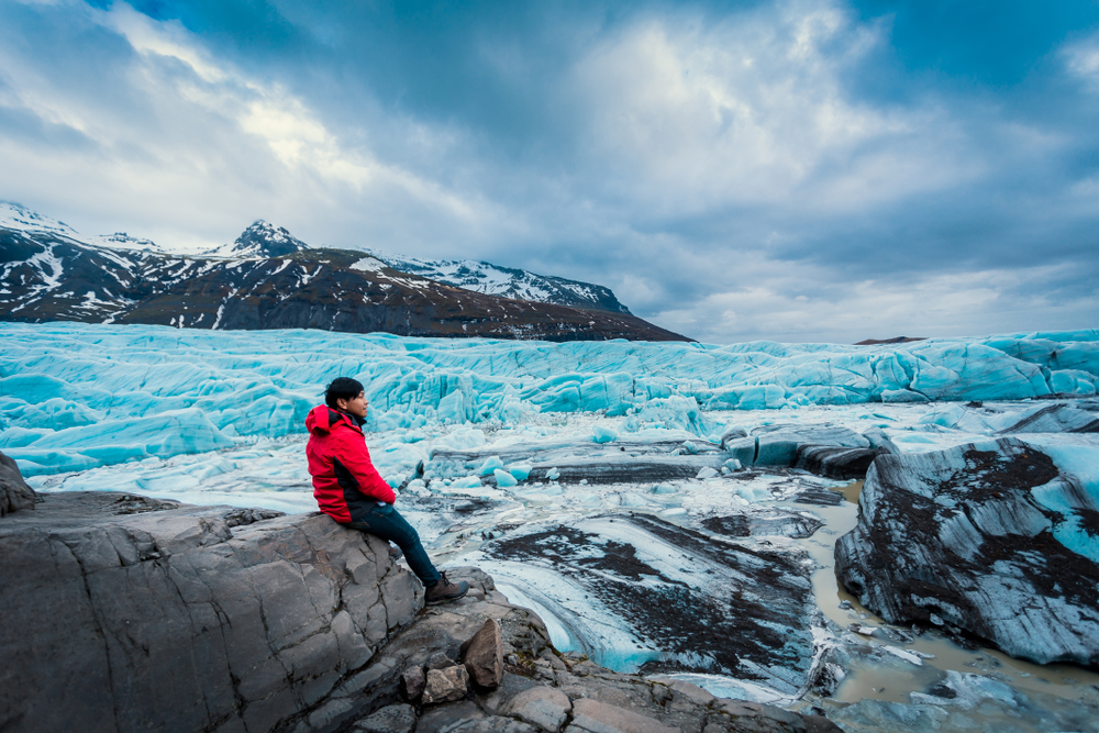 man in a red jacket and jeans sitting on a ledge looking out with a large glacier, mountains, and ice chunks behind him on a moody day