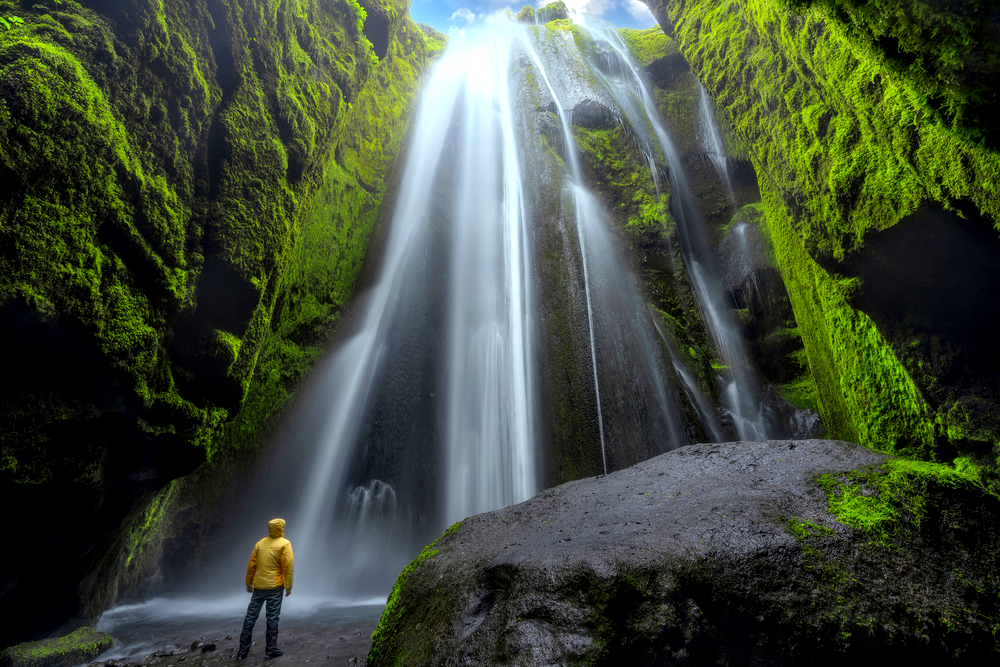 A person in a yellow jacket from their Iceland packing list looking up at a waterfall in a cave 
