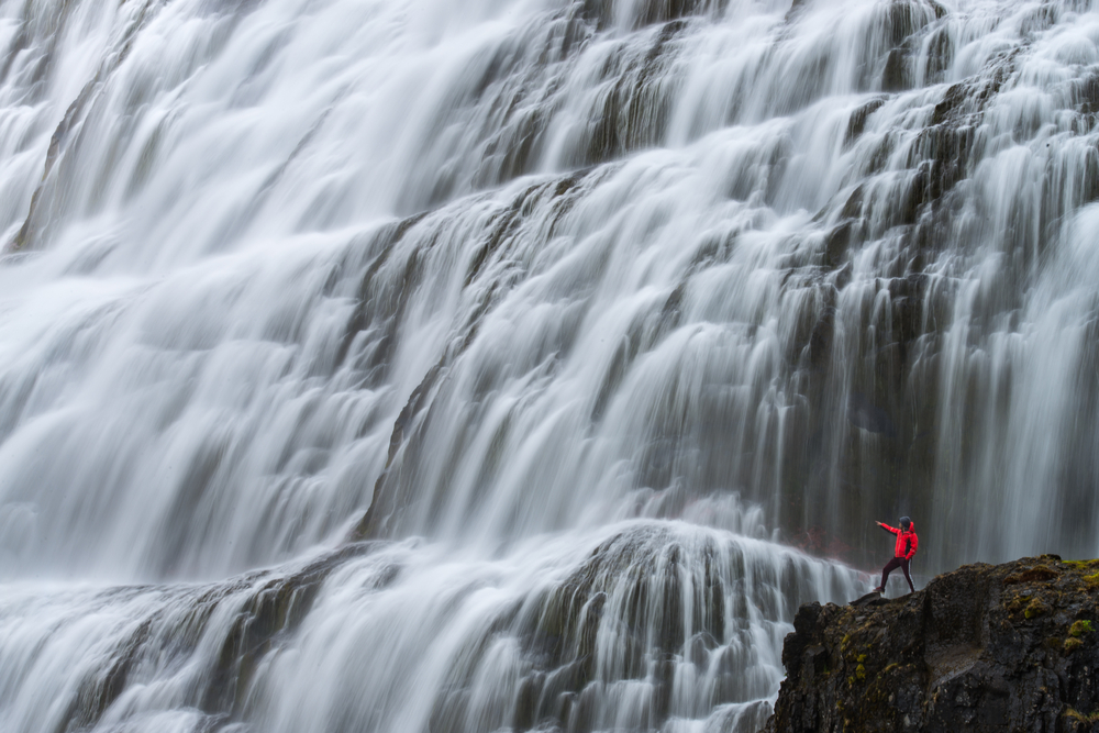 A person in a red coat on the side of a cliff pointing to a massive waterfall. The waterfall takes up most of the picture and is several layers of water cascading over massive rock formations. 