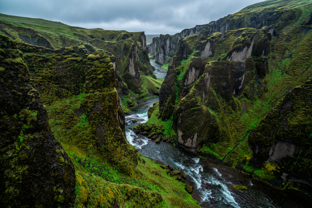 A large canyon made of large rock formations covered moss. There is a river rushing through the canyon over rocks. The sky is cloudy. 
