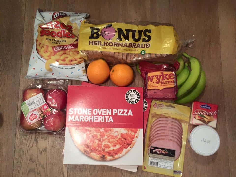 an assortment of groceries, bread, ham, frozen pizza, apples, chips, and fruits on a wood table