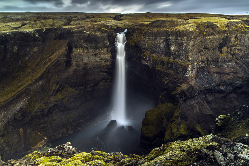 A large waterfall cascading down into a canyon. the canyon is made of dark volcanic rock and there is moss growing on the rocks. The waterfall has a few cascades before it falls directly into the canyon beneath it. One of the best stops on an Iceland road trip.