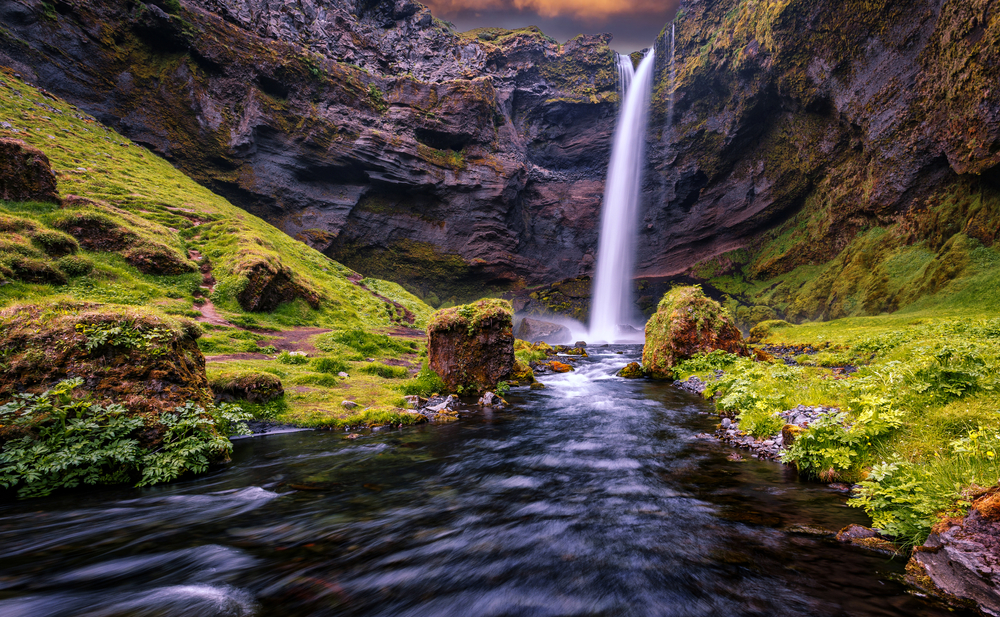 waterfall flowing into a river below surrounded by large rocks and green grass at sunset one of the best hiking trails in Iceland