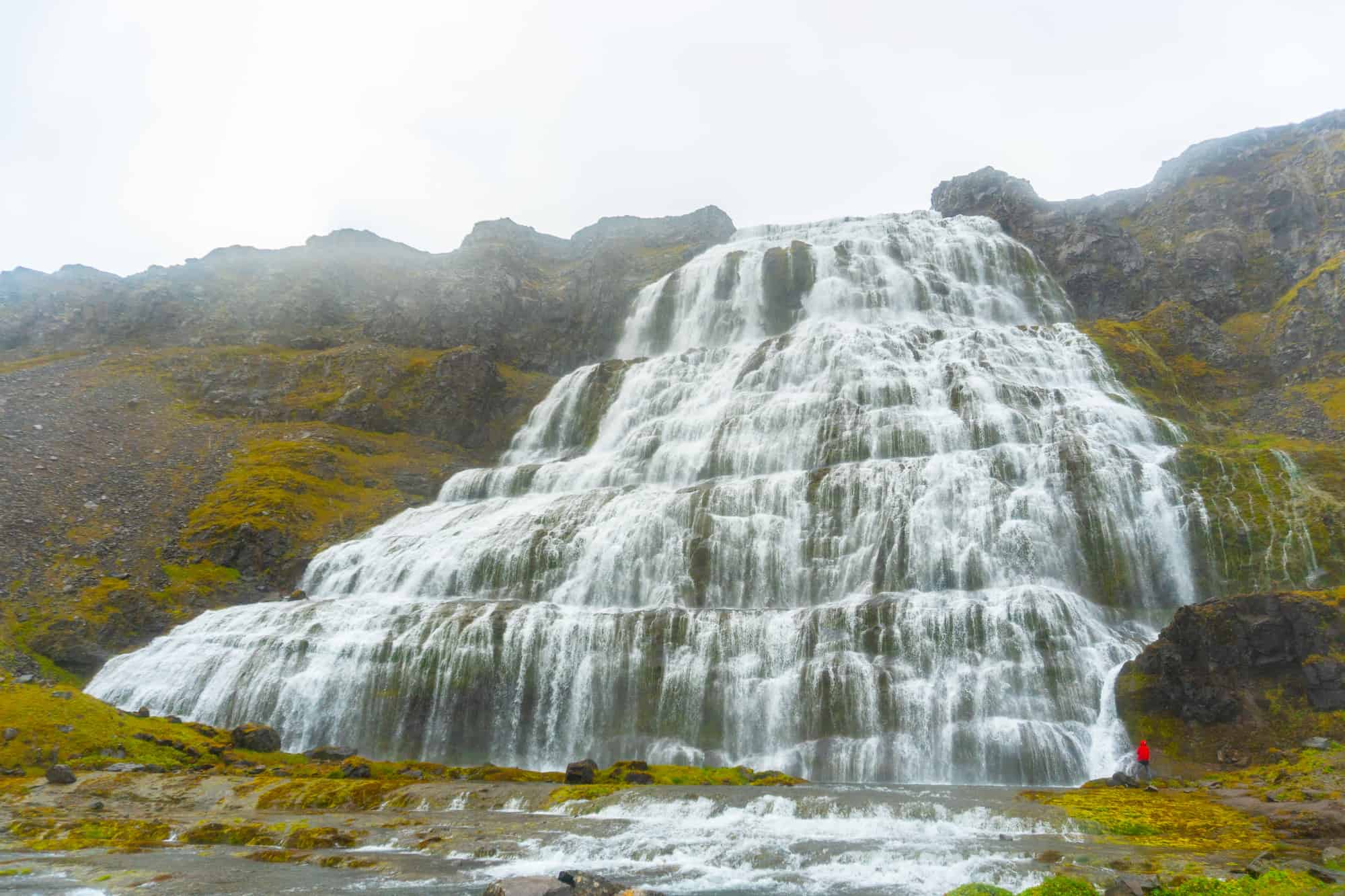 large waterfall on a cloudy day with a person in a red jacket looking up at it after one of the best hikes in iceland