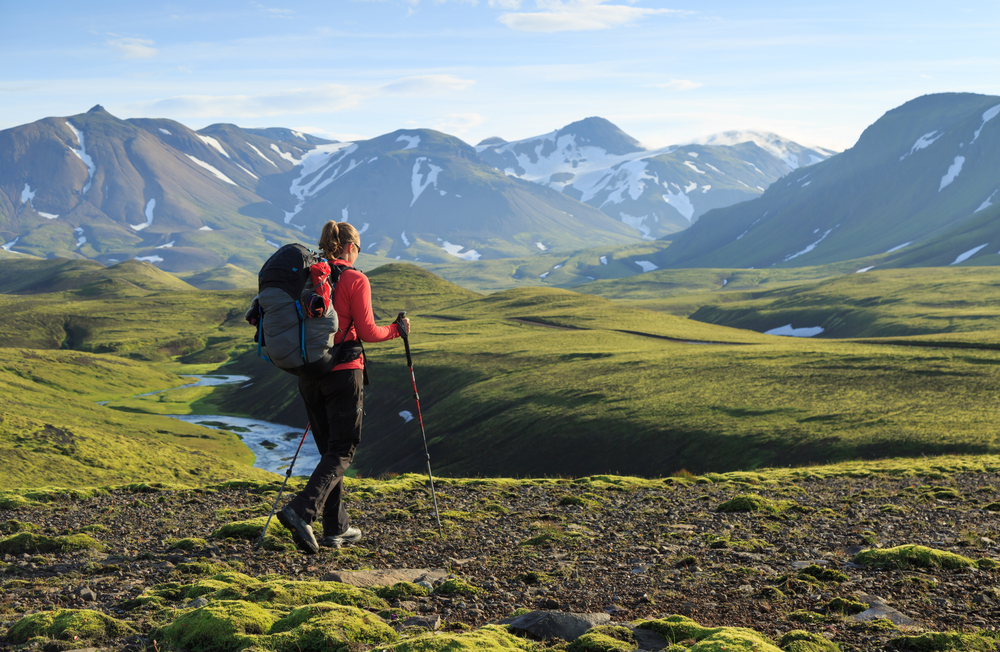 woman in a red jacket with a hiking bag and walking sticks. The landscape is mostly green and she's hiking in Iceland on a nice sunny day towards snow covered mountains