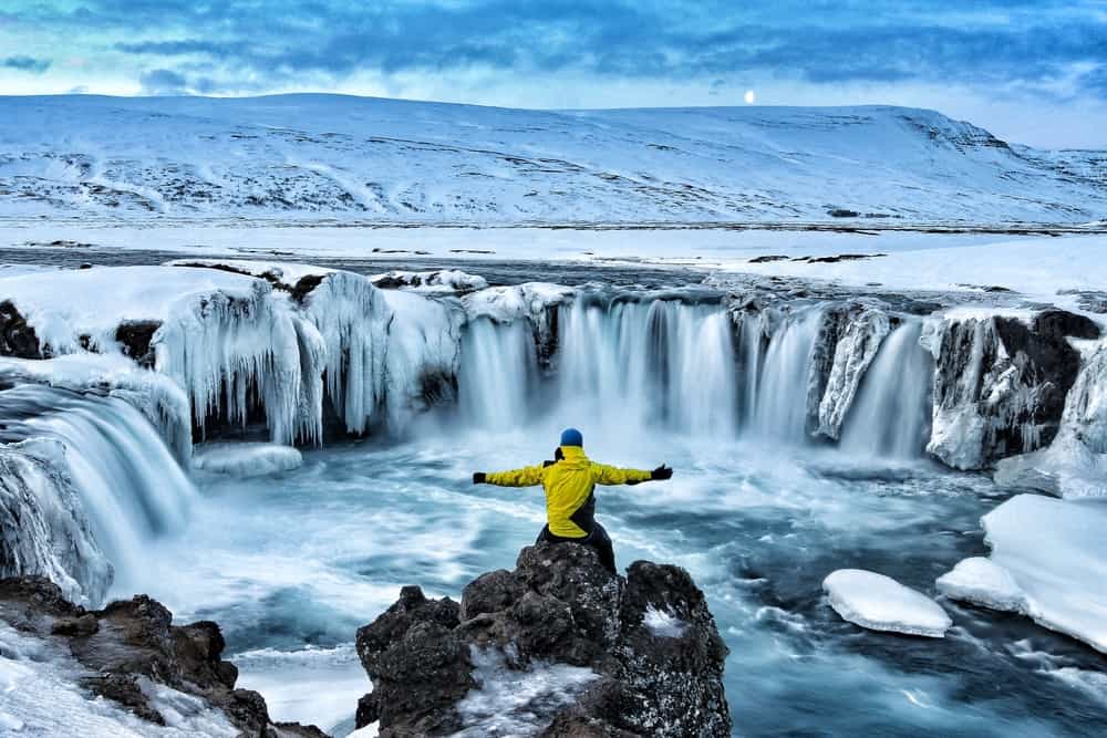 Mani n yellow jacket with his arms spread out sitting on a rock, looking at a waterfall surrounded by snow and ice in december in iceland