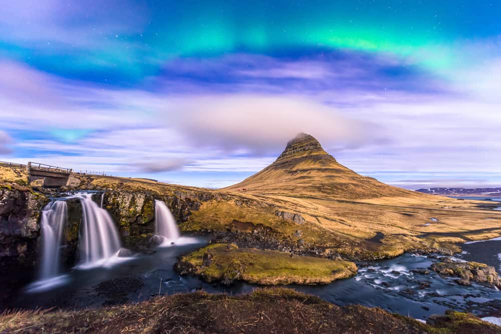 waterfall flowing into a river with a sloped mountain in the background and northern lights in the sky