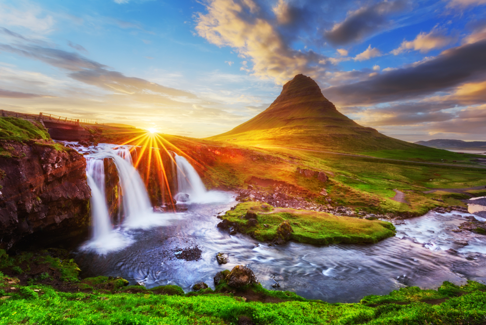 A mountain that looks like the point of a hat in the distance. In the front of a picture is a small waterfall that cascades into a river. There is grass and moss covering the mountain and the fields around it. 