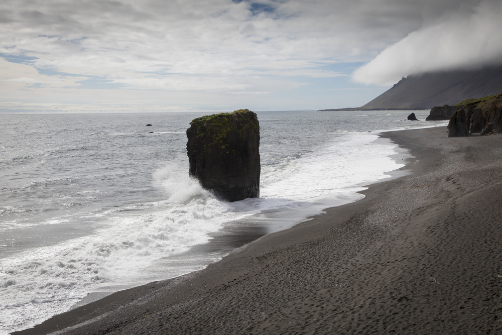 A black sand beach with rock formations on the beach and in the water. There is a large rectangular rock formation right where the waves crash that has moss on it. In the distance you can see the slope of a mountain. 