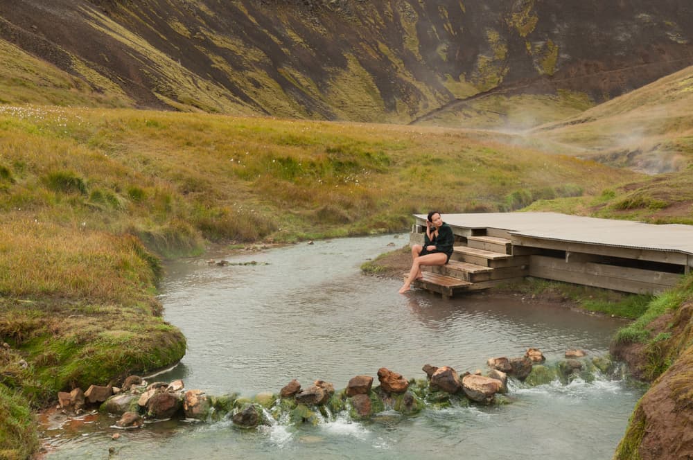 woman sits on stairs along river at Reykjadalur Hot Springs