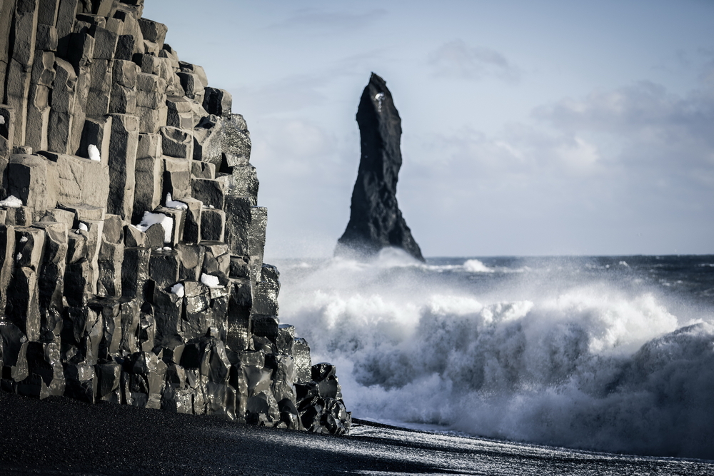 A black sand beach with a geometric rock formation on the beach. In the ocean you can see a large standing black rock formation. There are waves crashing on the shore. One of the best stops on an Iceland road trip.