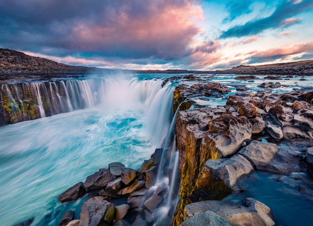 A large horse shoe shaped waterfall in Iceland. it is full of rocks and cascades over the entire edge of the horse shoe. It flows down into a river. The water is crystal blue and the sky is blue, pink, and purple with clouds. 