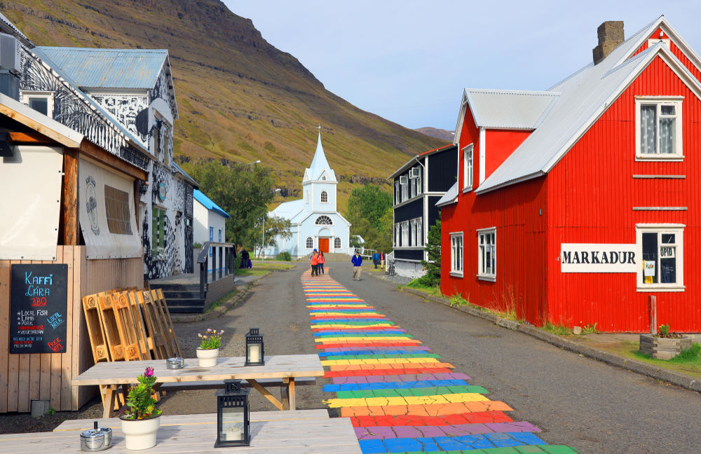 A street in a small town in Iceland. Down the street there is a rainbow painting, small shops, and a white church. Behind the church you can see the slope of a mountainside. 