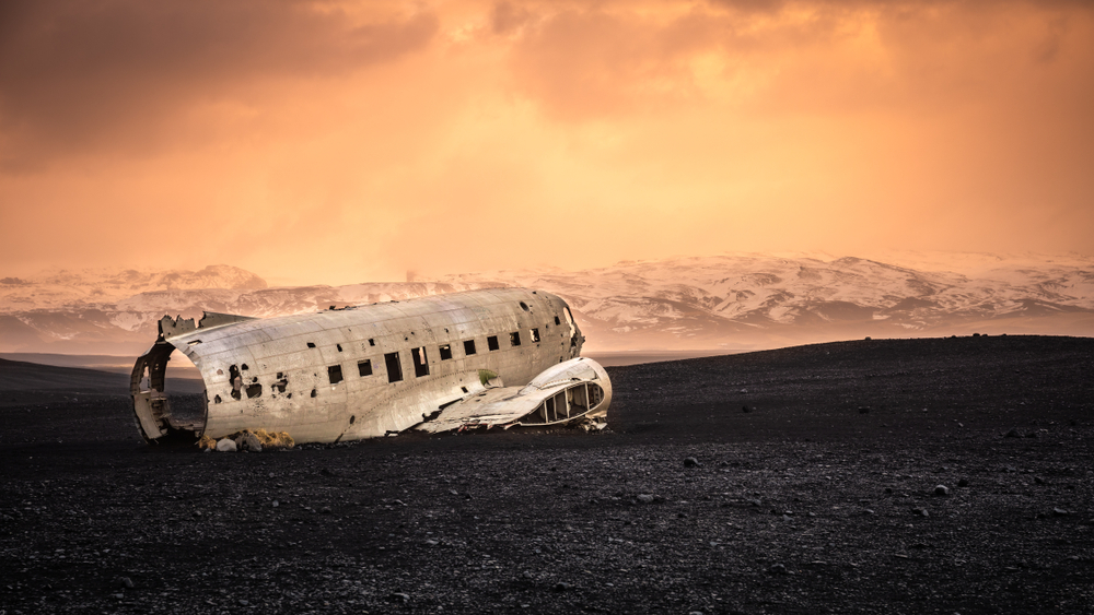 A hollowed out plane from a plane wreck on a black sand beach. In the distance you can see mountains covered in snow. The sky is a orange and yellow shade and looks hazy. 